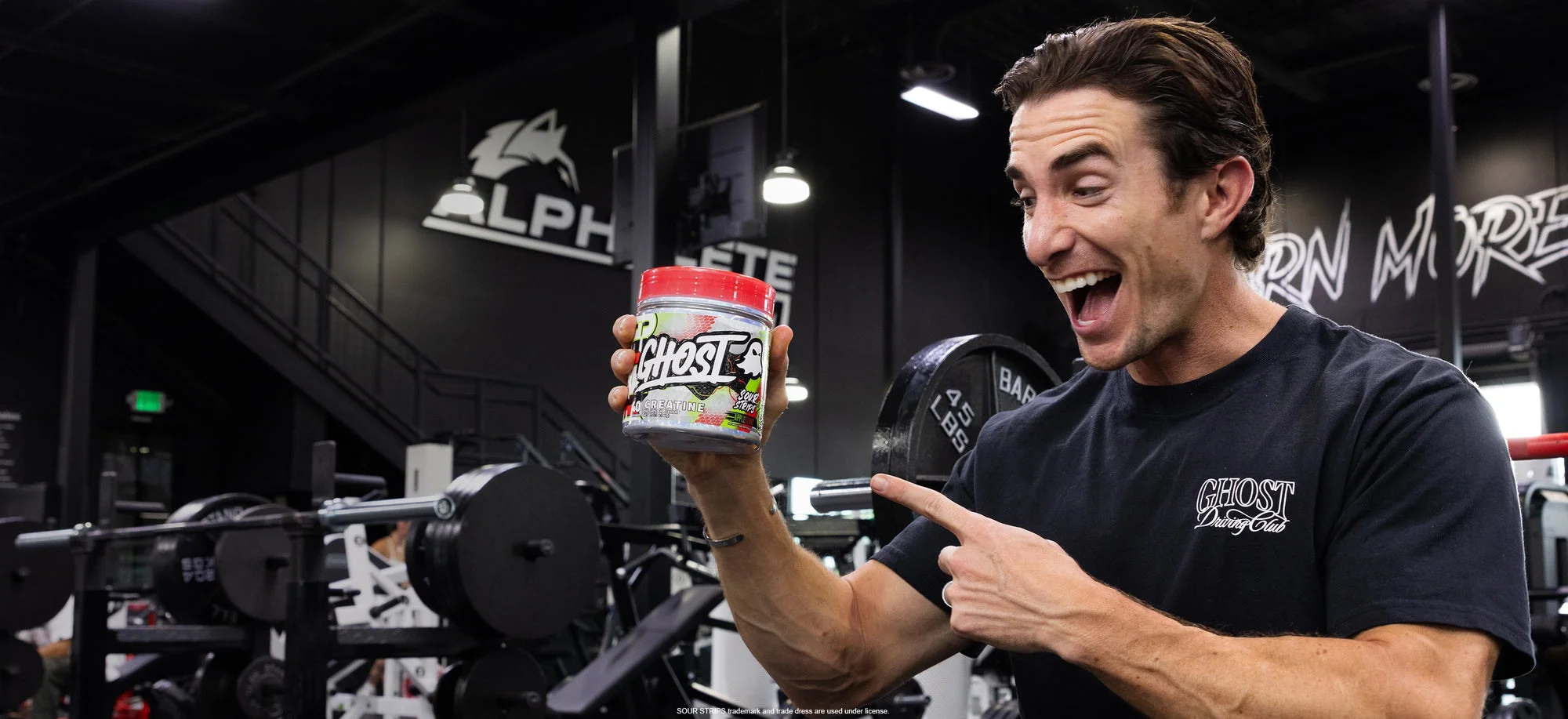 A GHOST® athlete in the gym pointing at a tub of GHOST® CREATINE x SOUR STRIPS® Appleberry
